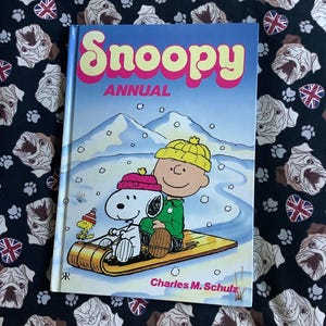 May include: A hardcover "Snoopy Annual" book featuring Snoopy and Charlie Brown sledding in a winter scene. The cover has the title in pink and yellow lettering. The background is a black fabric with pug dog and Union Jack patterns.