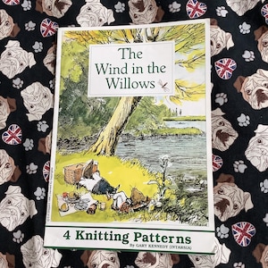 May include: A book titled "The Wind in the Willows" with an illustration of a picnic scene. The book cover is on a black fabric patterned with pug faces, paw prints, and Union Jack hearts. The book also includes "4 Knitting Patterns" text.