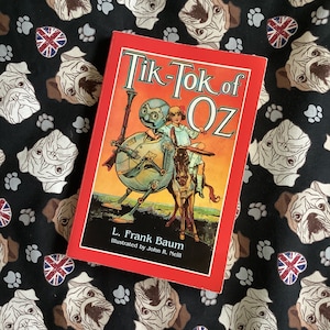 Może przedstawiać: Książka w twardej oprawie zatytułowana "Tik-Tok of Oz" autorstwa L. Franka Bauma, ilustrowana przez Johna R. Neilla. Okładka przedstawia kolorową ilustrację robota i dziewczynki. Książka leży na czarnej tkaninie z wzorem mopsów, śladów łap i serc z flagą brytyjską.