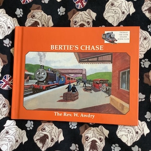 May include: A children's book with an orange cover, titled "Bertie's Chase". The cover illustration shows a blue train at a station. The book is part of the Thomas the Tank Engine Book Club. The author is The Rev. W. Awdry.
