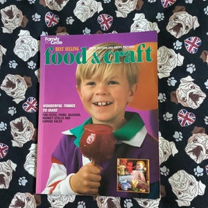 以下が含まれることがあります： 「food & craft」というタイトルの雑誌の表紙には、赤いリンゴ飴を持った笑顔の子供が写っています。背景は、白いブルドッグの顔、足跡、ユニオンジャックのハートが描かれた黒い生地です。雑誌はFamily Circleからです。