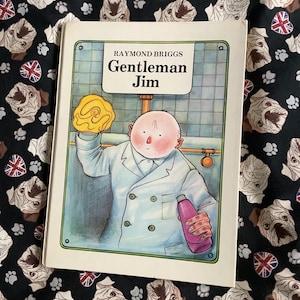 Puede incluir: Un libro titulado "Gentleman Jim" de Raymond Briggs. La portada muestra a un hombre con bata blanca con una esponja amarilla y una botella rosa. El libro est&aacute; sobre una tela negra con un estampado de pugs y banderas brit&aacute;nicas.