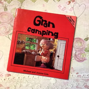 May include: A red book titled "Gran Camping" with a cartoon illustration of a character inside. The book has the text "As seen on BBC TV" and the authors' names, Michael and Joanne Cole. The book is on a floral and lace background.