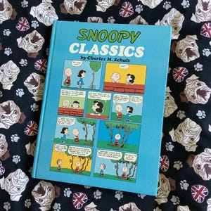 May include: A hardcover book titled "SNOOPY CLASSICS" by Charles M. Schulz. The book has a light blue cover with comic strip panels featuring Snoopy and other Peanuts characters. The book is on a black fabric with a pattern of pugs and Union Jack flags.