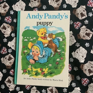 以下が含まれることがあります： 「Andy Pandy's puppy」というタイトルのヴィンテージ子供向けの本。子供と子犬のカラフルなイラストが描かれています。本は緑色の背表紙で、ブルドッグの顔と足跡がパターン化された黒い生地の上に置かれています。
