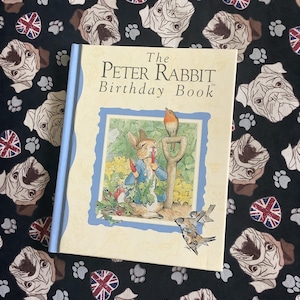 Può includere: Un libro con copertina rigida, "The Peter Rabbit Birthday Book", con dorso azzurro chiaro. La copertina presenta un'illustrazione di Peter Rabbit. Il libro è appoggiato su un tessuto nero con motivi di carlini, impronte di zampe e bandiere britanniche.