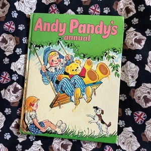 以下が含まれることがあります： 緑色の表紙とピンク色の文字が特徴の、ヴィンテージの「Andy Pandy's Annual」の本。表紙のイラストは、ブランコに乗った少年とテディベア、手前に女の子と犬が描かれています。背景は、白いブルドッグの顔とユニオンジャックの旗が描かれた黒い生地です。