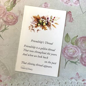 May include: A white card featuring the text "Friendship's Thread" and a poem about friendship. The card is adorned with a floral arrangement of brown, white, and purple flowers. The background has a floral pattern.