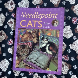 May include: A book titled "Needlepoint CATS" by Julie Hasler, featuring images of needlepoint cat portraits. The book is set against a dark background with a pattern of white dog faces, paw prints, and Union Jack flags.