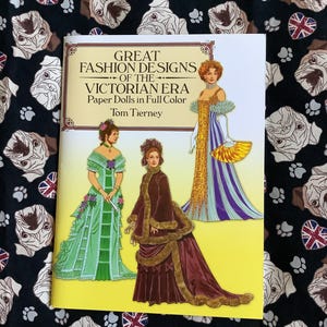 May include: A softcover book titled Great Fashion Designs of the Victorian Era Paper Dolls in Full Colour by Tom Tierney. The cover features three illustrations of figures in historical gowns, including a green dress, a brown coat, and a purple dress.