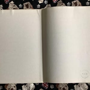 Puede incluir: Un libro abierto con p&aacute;ginas en blanco. Un peque&ntilde;o sello circular en relieve es visible en la p&aacute;gina derecha. El libro descansa sobre una tela negra estampada con caras de bulldog, huellas de patas y banderas de la Union Jack.