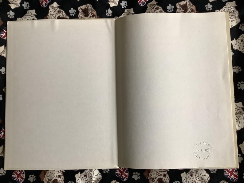 Puede incluir: Un libro abierto con p&aacute;ginas en blanco. Un peque&ntilde;o sello circular en relieve es visible en la p&aacute;gina derecha. El libro descansa sobre una tela negra estampada con caras de bulldog, huellas de patas y banderas de la Union Jack.