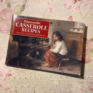 May include: A vintage cookbook titled "Favourite Casserole Recipes" with a red banner. The cover features a painting of a girl, a dog, and a kitchen scene. The book is on a floral and lace background.