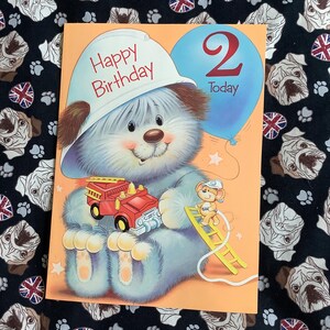 May include: A birthday card with a cartoon bear wearing a white firefighter's helmet that says "Happy Birthday." The bear holds a red fire truck. A blue balloon with the number "2" is also on the card. A mouse is climbing a yellow ladder.