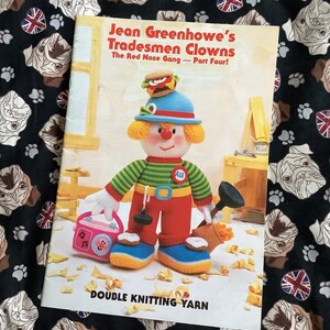 Puede incluir: Un libro titulado "Jean Greenhowe's Tradesmen Clowns" con una ilustración de payaso tejido con un sombrero de hamburguesa. El libro está sobre una tela negra estampada con caras de bulldog y banderas de la Union Jack. El texto "DOUBLE KNITTING YARN" es visible.