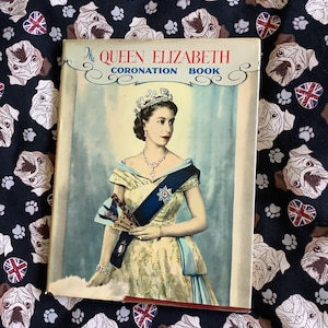 Puede incluir: Un libro antiguo titulado "The Queen Elizabeth Coronation Book" con un retrato de la reina Isabel II. El libro está sobre un fondo oscuro con un estampado de carlinos y banderas de la Union Jack.