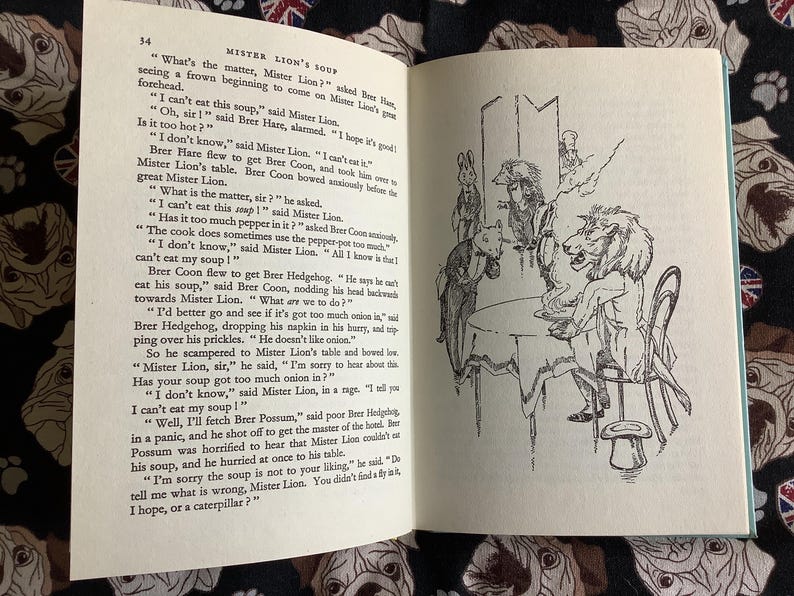 May include: An open book with a black and white illustration of a lion-headed character seated at a table with other animal figures. The text on the page reads "Mister Lion's Soup."
