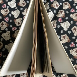 Puede incluir: Un libro abierto con p&aacute;ginas blanquecinas y una cubierta crema. El libro est&aacute; sobre un fondo negro estampado con caras de carlino, huellas de patas y banderas de la Union Jack. Las p&aacute;ginas est&aacute;n ligeramente amarillentas.