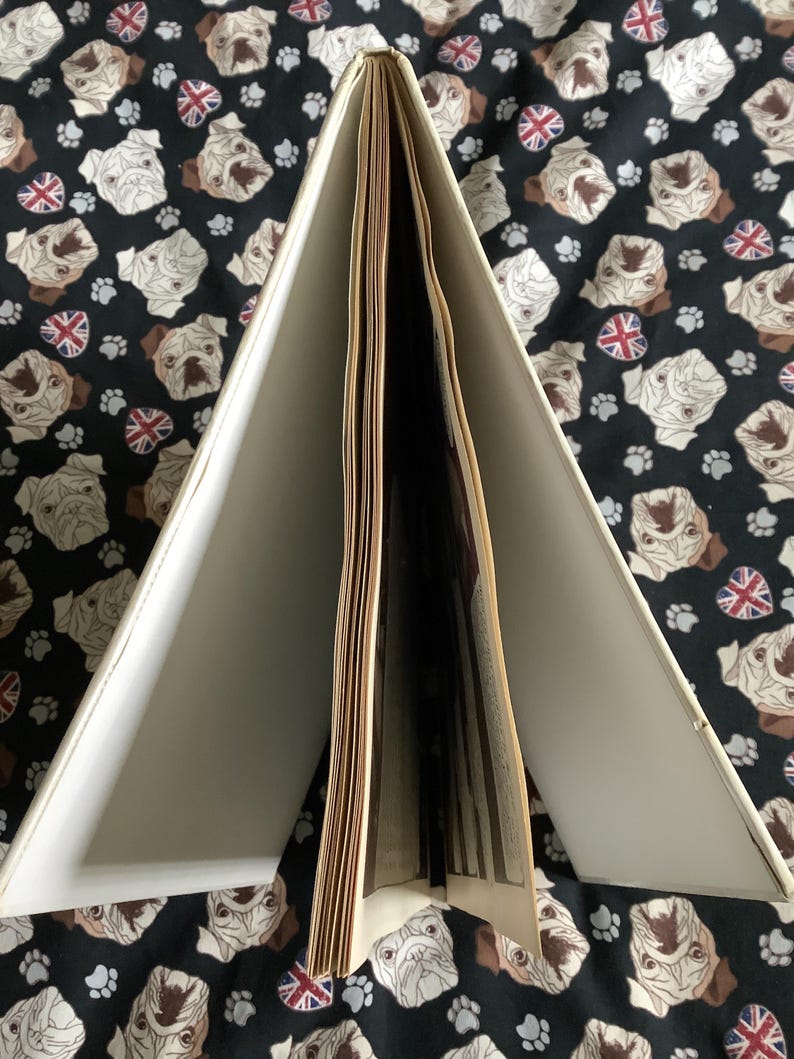 Puede incluir: Un libro abierto con p&aacute;ginas blanquecinas y una cubierta crema. El libro est&aacute; sobre un fondo negro estampado con caras de carlino, huellas de patas y banderas de la Union Jack. Las p&aacute;ginas est&aacute;n ligeramente amarillentas.