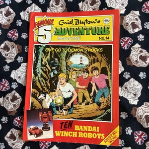May include: A vintage "Famous Five Adventure Magazine" No. 14, featuring a colorful illustration of children exploring a cave with a lighthouse in the distance. The cover also advertises "Ten Bandai Winch Robots". The magazine is set against a black fabric with a pug dog pattern.