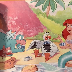 Pu&ograve; includere: Un'illustrazione di cartone animato di una sirena, un drago e un gabbiano che giocano a carte su una spiaggia. La sirena indossa un reggiseno di conchiglia e una coda verde. Il drago &egrave; verde e ha una pancia rosa. Il gabbiano &egrave; bianco con un becco giallo e zampe rosse.