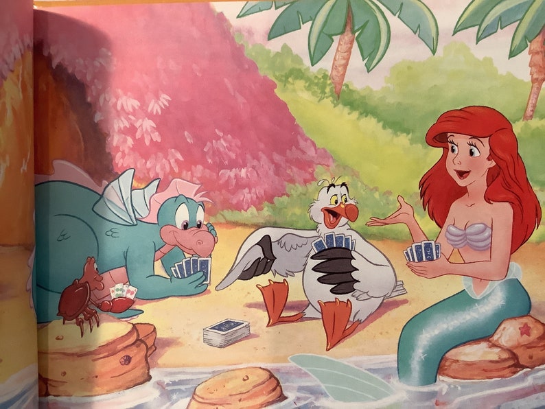 Pu&ograve; includere: Un'illustrazione di cartone animato di una sirena, un drago e un gabbiano che giocano a carte su una spiaggia. La sirena indossa un reggiseno di conchiglia e una coda verde. Il drago &egrave; verde e ha una pancia rosa. Il gabbiano &egrave; bianco con un becco giallo e zampe rosse.