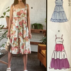 May include: A sewing pattern for a tiered dress with three different view options. The pattern includes instructions for making a dress with a floral print, a light blue dress, and a pink and purple dress.
