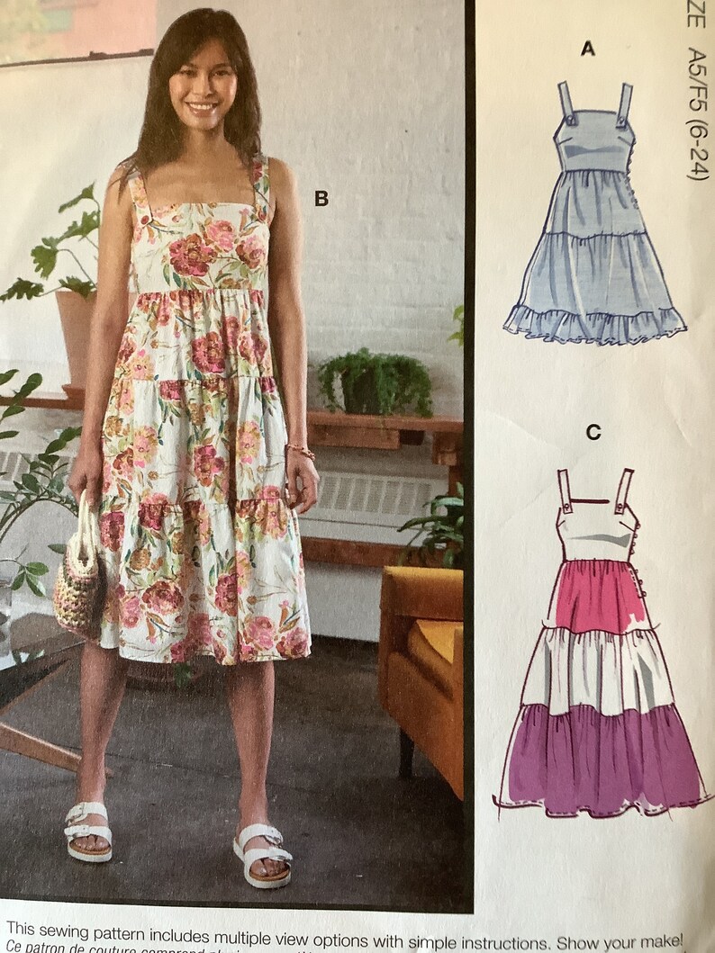 May include: A sewing pattern for a tiered dress with three different view options. The pattern includes instructions for making a dress with a floral print, a light blue dress, and a pink and purple dress.