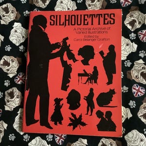 May include: A red book titled "Silhouettes: A Pictorial Archive of Varied Illustrations" edited by Carol Belanger Grafton. The cover features black silhouettes of people and objects.