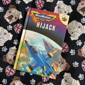 May include: A purple and blue book with the title "Micro Machines Hijack" on the cover. The cover features a blue and yellow jet plane flying over a globe. A small bee is in the top right corner.