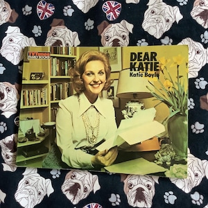 May include: A vintage book cover titled "Dear Katie" by Katie Boyle, featuring a smiling woman in a white blouse. The background is a dark fabric patterned with bulldog faces, paw prints, and Union Jack emblems.