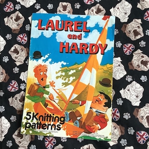 May include: A vintage book cover featuring Laurel and Hardy in a cartoon illustration. The book is titled "Laurel and Hardy 5 Knitting Patterns by Gary Kennedy (Intarsia)".