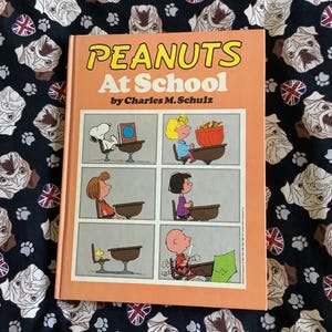 May include: A hardcover book titled "PEANUTS At School" by Charles M. Schulz. The cover is orange with the title in yellow and the book features comic strip panels of Peanuts characters. The book is on a black background with a pattern of pugs and Union Jack flags.