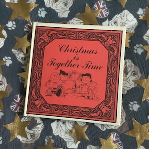 May include: A square, red box with the text "Christmas is Together-Time" and a cartoon illustration of the Peanuts gang. The box is on a blue fabric with gold stars and dog faces.