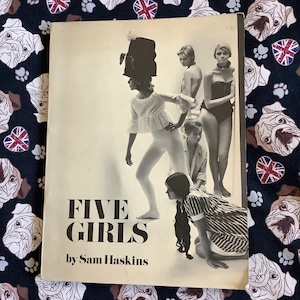 May include: A vintage photography book titled Five Girls by Sam Haskins with a cream-colored cover featuring a black and white image of five individuals in various poses, including one wearing a tall hat, set against a dark fabric background.