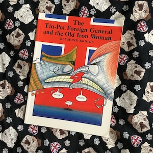 May include: A book titled "The Tin-Pot Foreign General and the Old Iron Woman" by Raymond Briggs. The cover features a colorful illustration of two figures in a battle scene with the words "Mine!" and "Help!" in speech bubbles.