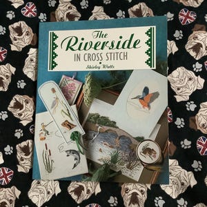 以下が含まれることがあります： Shirley Wattsの「The Riverside in Cross Stitch」というタイトルの本。川の風景や野生生物のクロスステッチパターンが特徴です。ブルドッグの顔、足跡、ユニオンジャックの旗が描かれた黒い生地の上に本が置かれています。