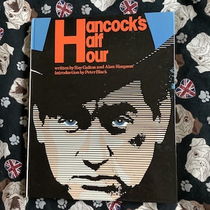 May include: A book titled "Hancock's Half Hour" with a graphic illustration of a man's face on the cover. The title is in orange and white, set against a black background with a blue border. The book is on a patterned fabric.