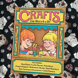 Könnte beinhalten: Ein Vintage-Buchcover mit dem Titel "CRAFTS FOR AGES 8 & UP" in rosa und gelben Buchstaben. Das Cover zeigt eine farbenfrohe Illustration von zwei Kindern, die durch ein Fenster ein Puppenspiel betrachten. Das Buch listet Bastelideen auf, darunter Pompons, Bastelspulen, Filzideen, Fingerpuppen, Halloween-Masken, Staubmonster, Filz-Weihnachtsornamente und Stoffblumen.