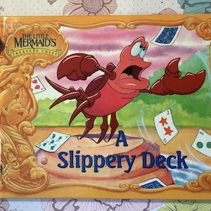 Pu&ograve; includere: Una copertina di libro che mostra un granchio rosso con un'espressione sorpresa su uno sfondo giallo. Il titolo blu dice "A Slippery Deck". Il granchio tiene una carta da gioco nella sua chela destra. La copertina del libro &egrave; della serie "The Little Mermaid's Treasure Chest".