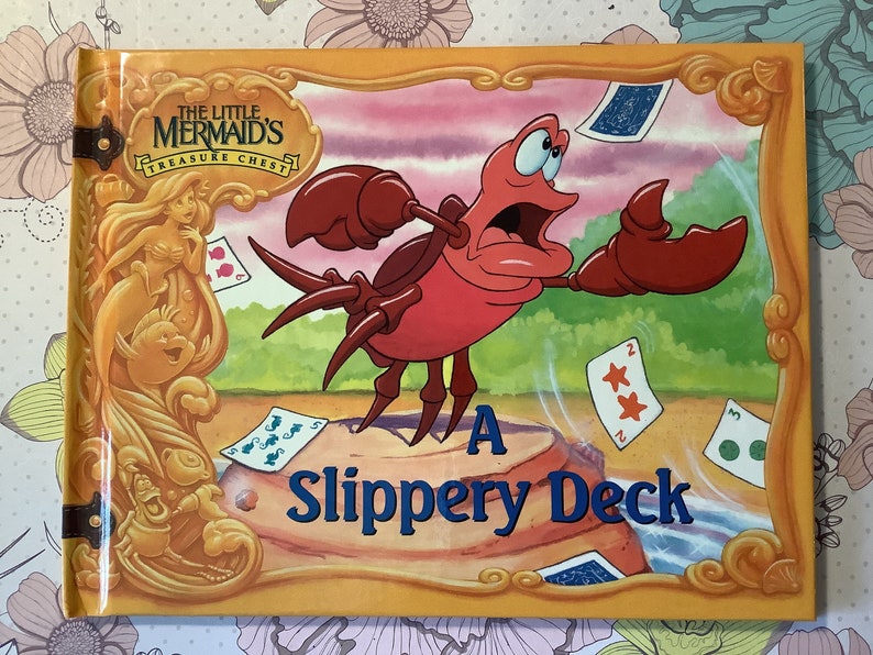 Pu&ograve; includere: Una copertina di libro che mostra un granchio rosso con un'espressione sorpresa su uno sfondo giallo. Il titolo blu dice "A Slippery Deck". Il granchio tiene una carta da gioco nella sua chela destra. La copertina del libro &egrave; della serie "The Little Mermaid's Treasure Chest".