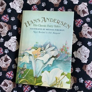 May include: A hardcover book titled "Hans Andersen His Classic Fairy Tales" with illustrations by Michael Foreman. The cover art features a fairy tale scene with a white flower and a castle. The book rests on a black fabric with pug dog and Union Jack patterns.