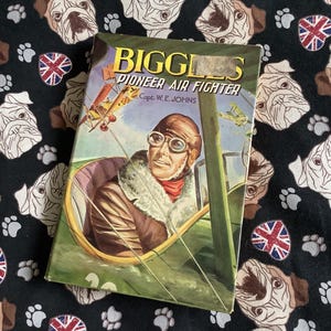 May include: A vintage book titled "Biggles Pioneer Air Fighter" by Capt. W.E. Johns. The cover features a colourful illustration of a pilot in the cockpit of a biplane.