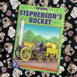 Könnte beinhalten: Ein grünes und gelbes Buch mit dem Titel "Bauen Sie Ihre eigene Stephenson's Rocket" und einem Bild einer gelben Dampflokomotive. Das Buch liegt auf einem schwarzen Stoff mit einem Muster aus Mops.