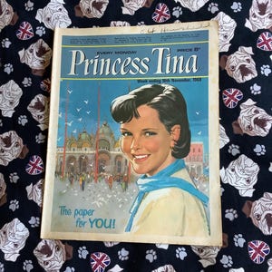 以下が含まれることがあります： 1968年11月のヴィンテージコミック「プリンセス・ティナ」で、表紙には笑顔の女性が描かれています。背景は青い空と都市景観です。コミックは、白いブルドッグの顔とユニオンジャックの旗が描かれた黒い生地の上にあります。