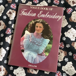 Könnte beinhalten: Ein burgunderfarbenes Buch mit dem Titel "Vogue Book of Fashion Embroidery" mit einem Foto einer Frau in einem weißen und rosa Kleid mit einem floral bestickten Ausschnitt. Das Buch wurde von Collins herausgegeben.