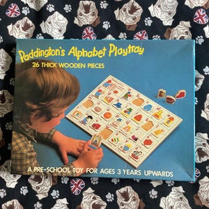 Peut inclure: Boîte de jeu Paddington's Alphabet Playtray avec 26 pièces en bois épaisses. La boîte bleue montre un enfant plaçant une lettre dans un plateau en bois avec des emplacements alphabétiques. Texte : Paddington's Alphabet Playtray, 26 THICK WOODEN PIECES, A PRE-SCHOOL TOY FOR AGES 3 YEARS UPWARDS.