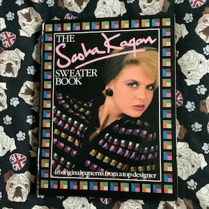 May include: A hardback book titled "The Sasha Kagan Sweater Book" with a woman on the cover. The cover has a colourful border and the text "48 original patterns from a top designer". The book is on a black fabric with bulldog and Union Jack patterns.