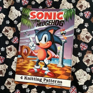 May include: A book titled "SONIC THE HEDGEHOG" featuring the character Sonic, with the text "4 Knitting Patterns". The book is on a black fabric patterned with pugs, paw prints, and Union Jacks.