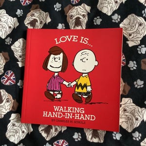 May include: A red book titled "LOVE IS... WALKING HAND-IN-HAND" by Charles M. Schulz. The cover features a cartoon couple holding hands. The book rests on a black fabric patterned with bulldog faces, paw prints, and Union Jack hearts.